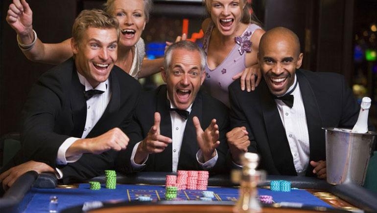 Мужчины в костюмах и женщины в вечерних платьях радуются за игорным столом успешной ставке