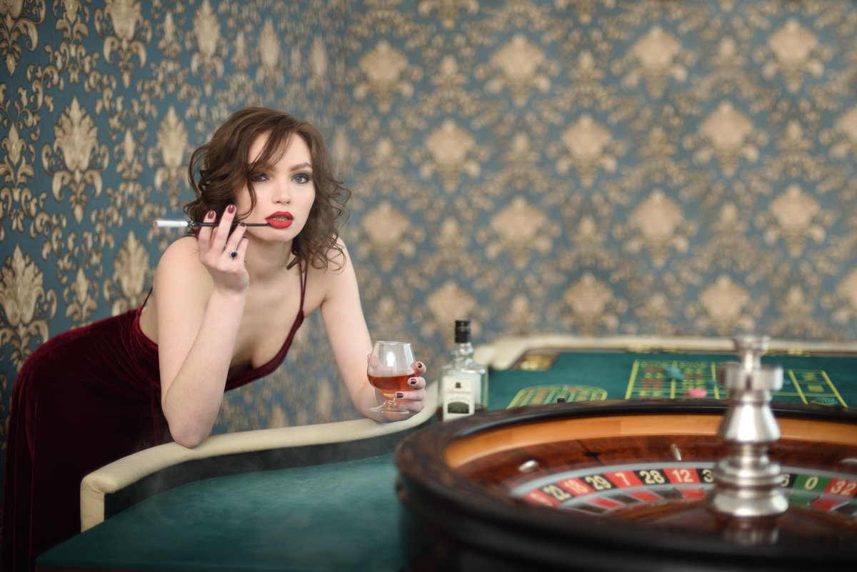 Красотка курит сигарету и распивает виски у стола для игры в рулетку
